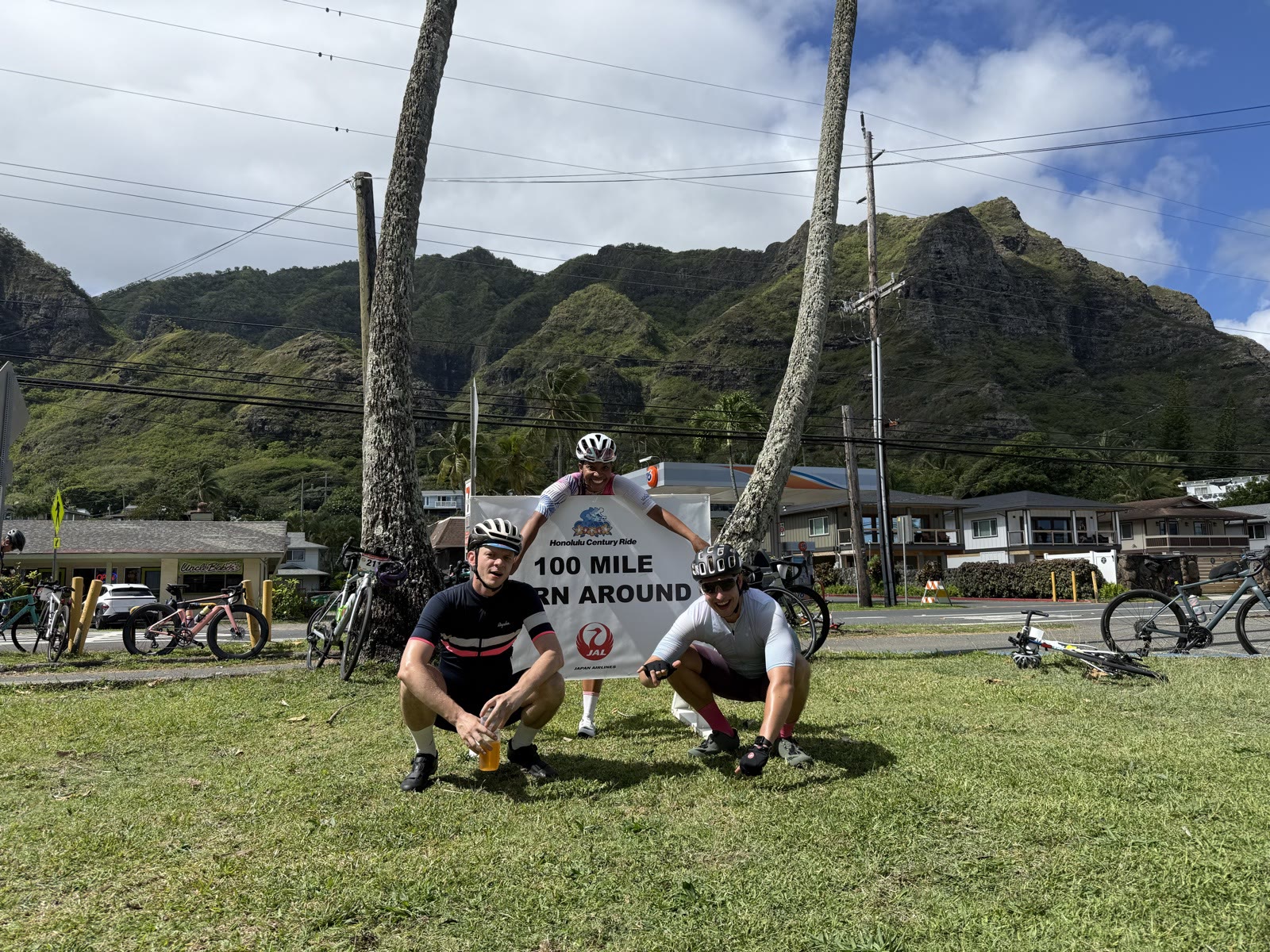 Three cyclists posing with the Honolulu Century Ride 100 mile turnaround sign on a grassy verge, with green mountains behind
