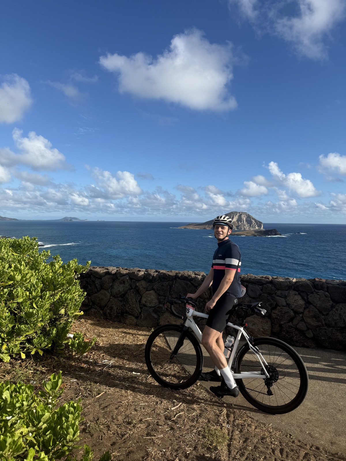 Pavel Bushuyeu in cycling kit and helmet standing with a white road bike at a seaside lookout, with the ocean and a headland behind