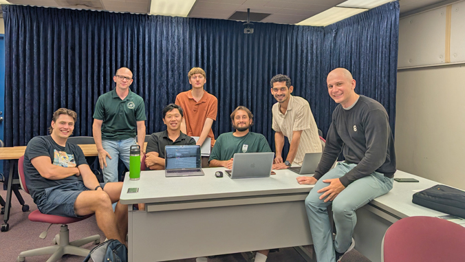 Seven members of the UH Mānoa team that won the Data Curation for Vision Language Reasoning challenge, seated and standing around a desk with laptops in front of a blue curtain