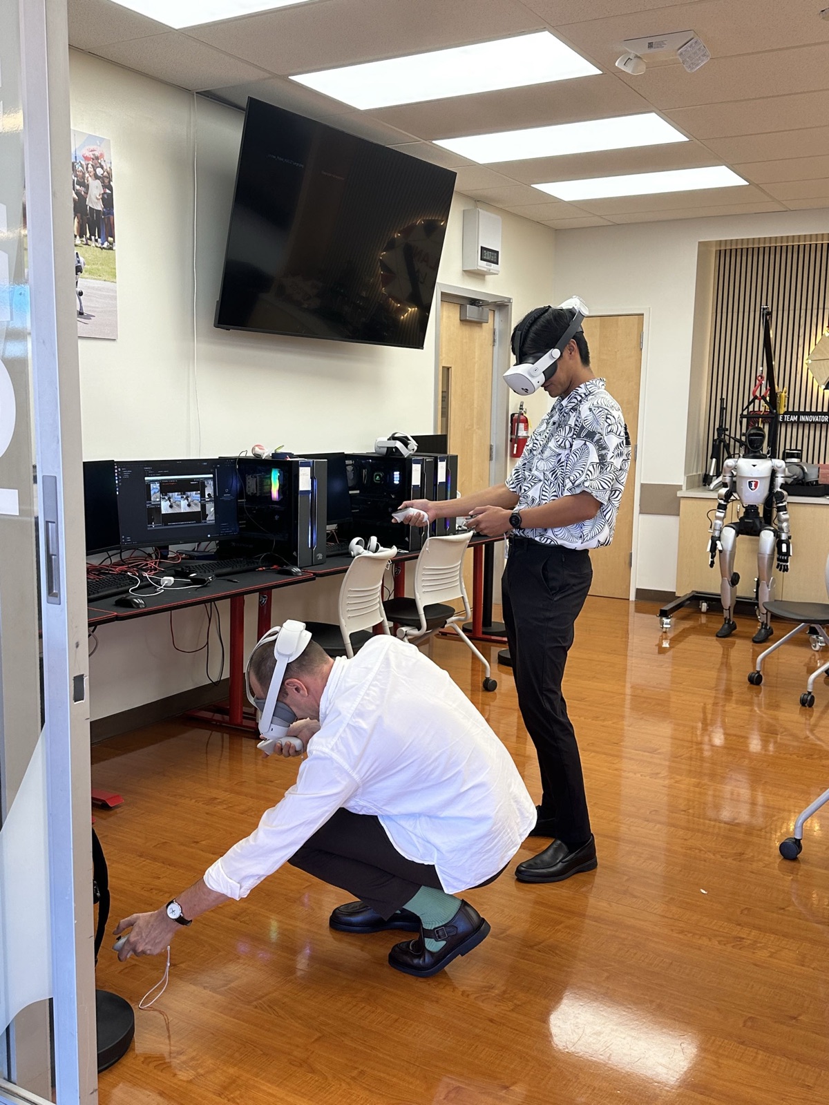 Two people in VR headsets with hand controllers in a robotics lab, one standing and one crouching, with a humanoid robot standing in the background