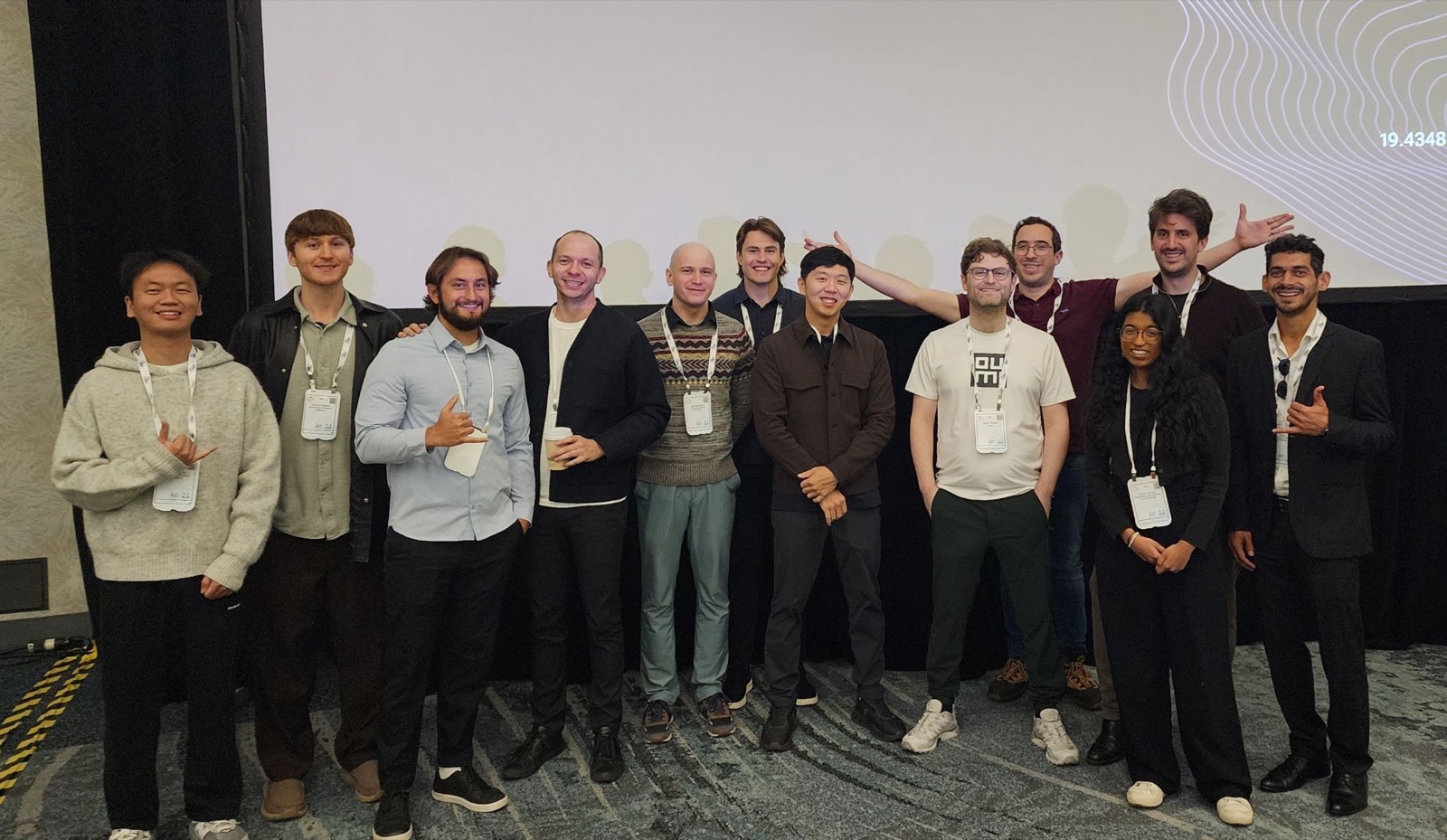 Twelve people, including the UH Mānoa team, posing in front of a projection screen at the DCVLR challenge session at NeurIPS 2025 in San Diego