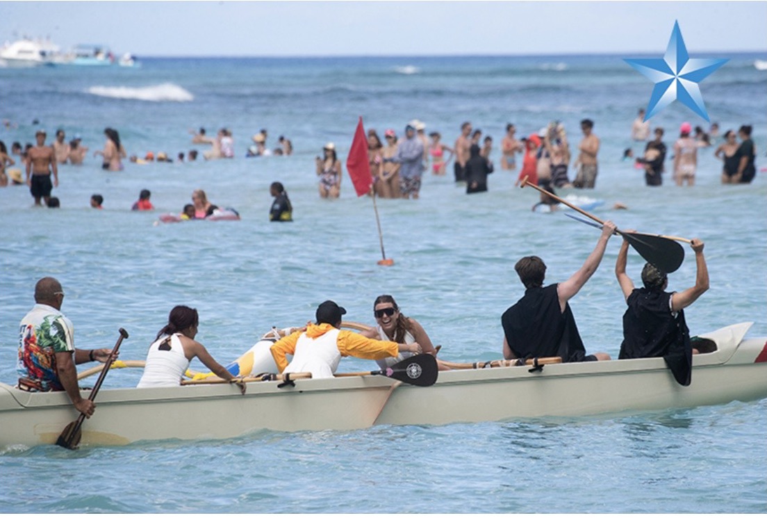 A mixed Hui Lanakila crew celebrating in their outrigger canoe at the finish of the Walter J. Macfarlane Regatta at Waikīkī Beach, paddles raised, with swimmers in the water behind